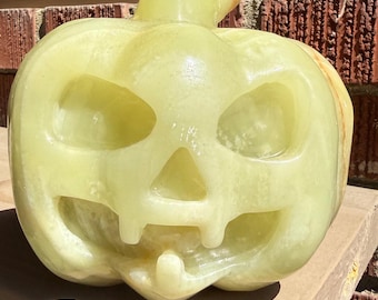 May include: A carved green onyx pumpkin sculpture featuring a jack-o'-lantern face with triangular eyes, a triangular nose, and a mouth with two teeth. The text reads: HQ Green Onyx Pumpkin, 12.5 lbs, 7x5.5” tall. The stone has a smooth, polished finish.