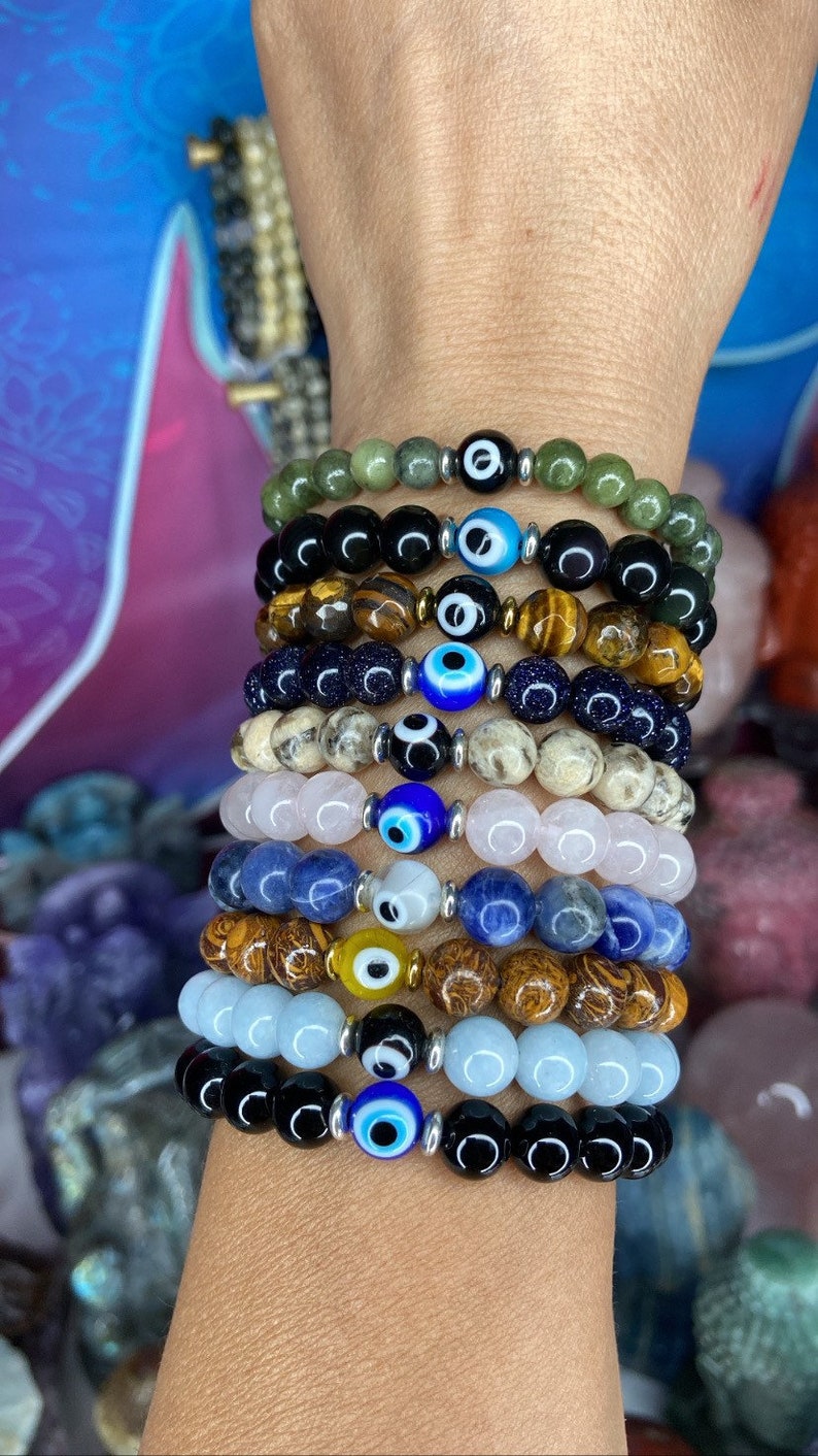 K&ouml;nnte beinhalten: Ein Stapel von acht Armb&auml;ndern mit verschiedenen farbigen Perlen und einem blauen b&ouml;sen Blick-Anh&auml;nger an jedem. Die Armb&auml;nder sind aus schwarzem Onyx, Tigerauge, blauem Lapislazuli, Rosenquarz, Aquamarin und anderen Steinen.
