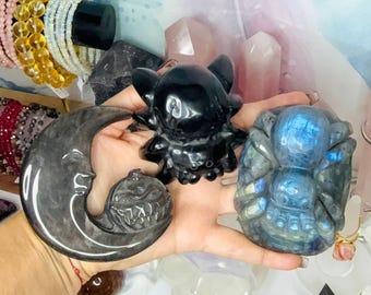 May include: A black obsidian crescent moon carving with a pumpkin face, a black obsidian cat figurine, and a blue iridescent labradorite spider carving. These three polished stone carvings are held together in a palm, showcasing detailed textures.