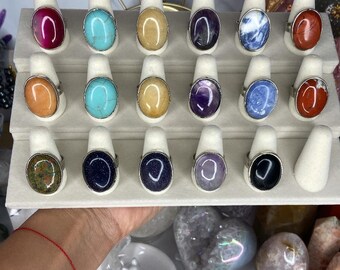 May include: A display of 18 oval-shaped gemstone rings on white stands. The rings feature a variety of colors, including red, blue, green, purple, and yellow.