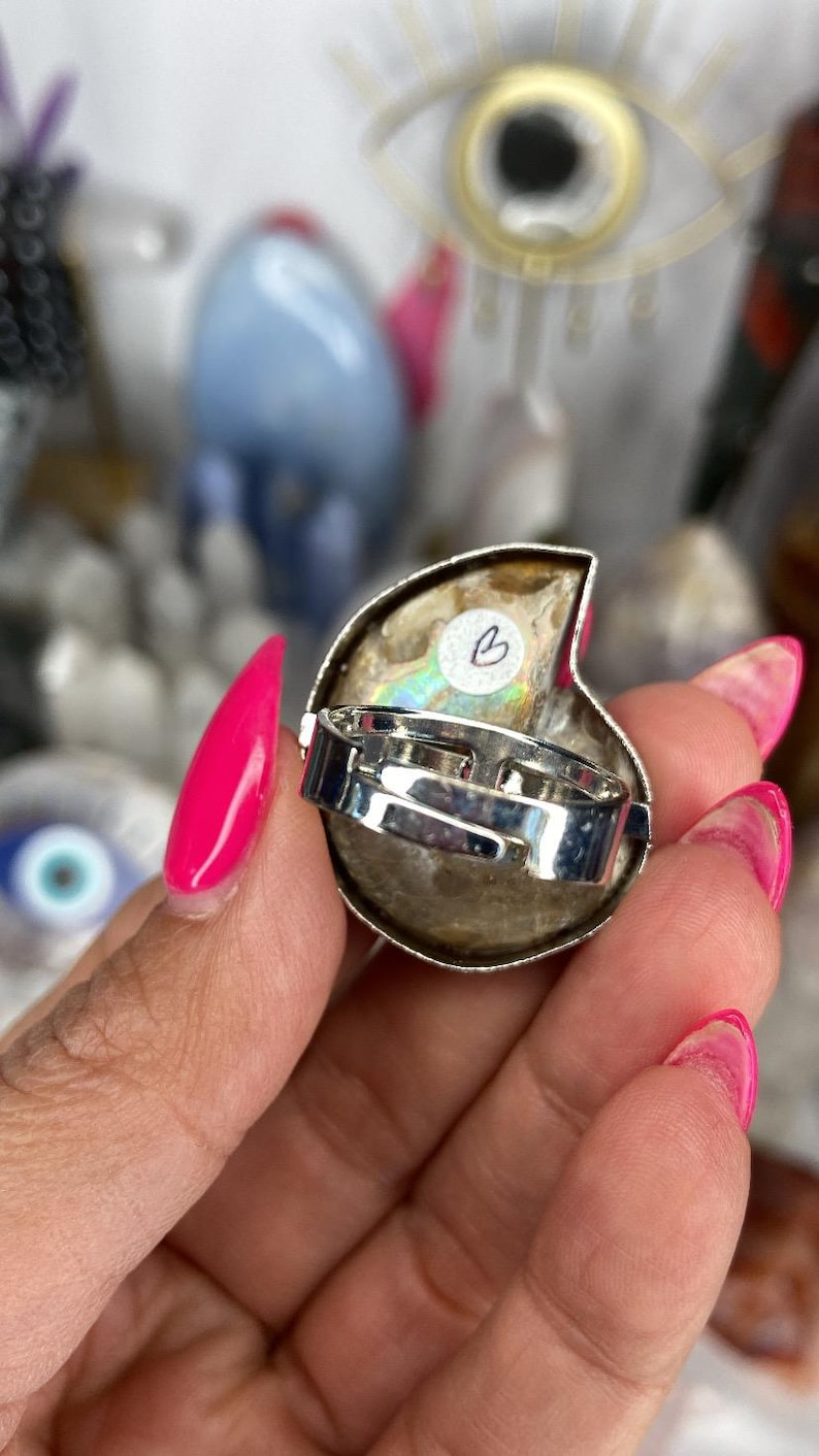 May include: A silver ring with a large iridescent ammonite stone. The stone is a light brown color with a pearly sheen. The ring is adjustable and has a simple design.