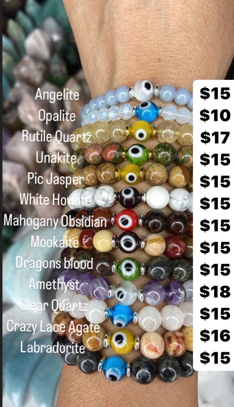 K&ouml;nnte beinhalten: Ein Stapel von 12 Armb&auml;ndern mit verschiedenen farbigen Steinen und einem b&ouml;sen Blick-Anh&auml;nger. Die Steine umfassen Angelit, Opalit, Rutilquarz, Unakit, Pic-Jaspis, wei&szlig;er Howlith, Mahagoni-Obsidian, Mookaite, Drachenblut, Amethyst, klarer Quarz, verr&uuml;ckte Spitzenachat und Labradorit. Die Armb&auml;nder sind zwischen 8 und 15 Euro erh&auml;ltlich.