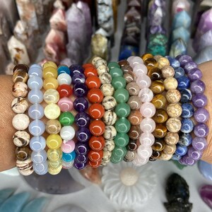 K&ouml;nnte beinhalten: Ein Stapel von 12 bunten Perlenarmb&auml;ndern aus verschiedenen nat&uuml;rlichen Steinen, darunter Amethyst, Tigerauge und Rosenquarz. Die Armb&auml;nder sind auf dem Handgelenk einer Person gestapelt.