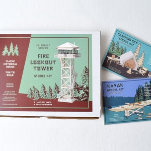 May include: A white box with a red and green design featuring a model kit of a fire lookout tower. The box includes the text "U.S. Forest Service Fire Lookout Tower Model Kit".  The box is next to two smaller boxes, one blue and one green, with model kits of a kayak and a camping set.