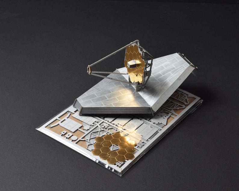 May include: A model of the James Webb Space Telescope, a large, gold-plated telescope with a hexagonal mirror, is shown on a white background. The model is made of metal and is assembled on a base.