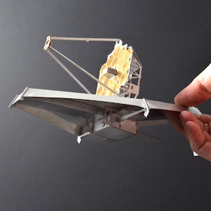 May include: A model of the James Webb Space Telescope, a large, silver, and gold space telescope with a hexagonal mirror, is held in a hand against a dark gray background.