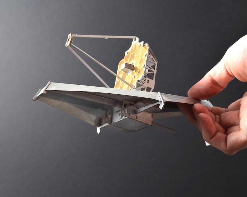 May include: A model of the James Webb Space Telescope, a large, silver, and gold space telescope with a hexagonal mirror, is held in a hand against a dark gray background.