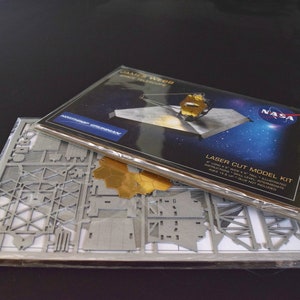 May include: A laser-cut model kit of the James Webb Space Telescope. The kit includes a detailed model of the telescope, along with instructions and a sheet of gold-colored metal.