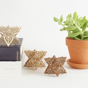 May include: Three wooden geometric star ornaments with intricate laser cut designs. The stars are arranged on a white surface with a potted succulent plant in the background.