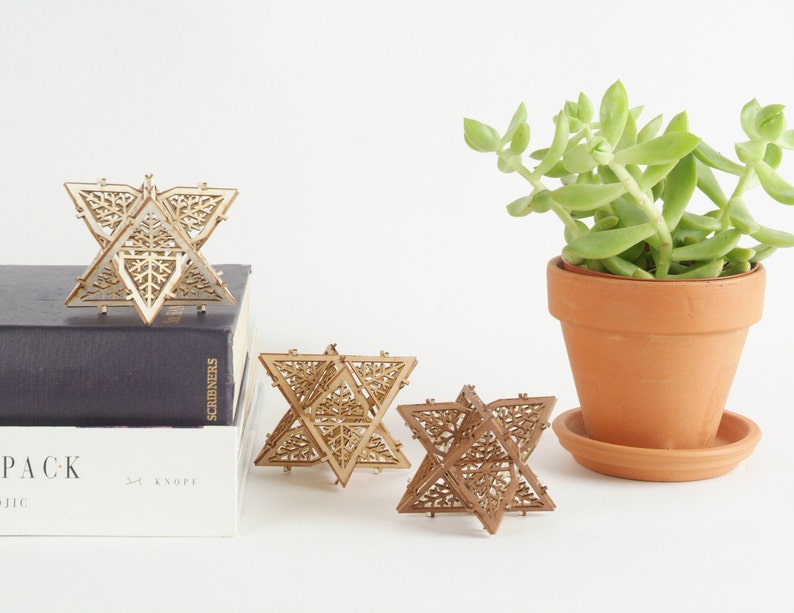 May include: Three wooden geometric star ornaments with intricate laser cut designs. The stars are arranged on a white surface with a potted succulent plant in the background.