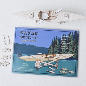 May include: A wooden kayak model kit with laser cut parts. The kit includes a kayak, kayak rack, paddle, backup paddle, life vest, bilge pump, and fishing pole. The box has a blue background with a lake scene and green trees. The text on the box reads "Kayak Model Kit Laser Cut Parts" and "Kit Includes: Kayak, Kayak Rack, Paddle, Backup Paddle, Life Vest, Bilge Pump, Fishing Pole".