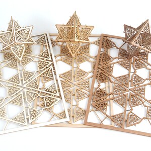 May include: Three unfinished wooden star-shaped ornaments with intricate laser-cut designs. The ornaments are layered on top of each other, with the largest ornament at the bottom and the smallest ornament at the top. The ornaments are made of light brown wood.