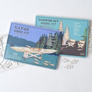 May include: Two laser cut wood model kits, one of a kayak and one of a camping set. The kayak kit includes a kayak, paddle, life preserver, fishing pole, and fish. The camping kit includes a tent, two chairs, a table, a campfire, and a tree. Both kits are on a white surface.