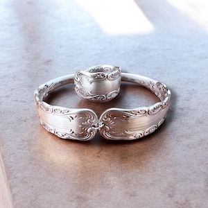 May include: A silver bracelet and ring set. The bracelet is made from two spoon handles connected by a hinge. The ring is made from a spoon bowl. Both pieces have ornate, engraved floral patterns.