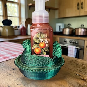 以下が含まれることがあります： 緑色の鶏の形をした石鹸ホルダーと石鹸のボトル。石鹸のボトルには、オレンジと白い花の画像が描かれたラベルが付いています。ホルダーは木製の表面にあります。