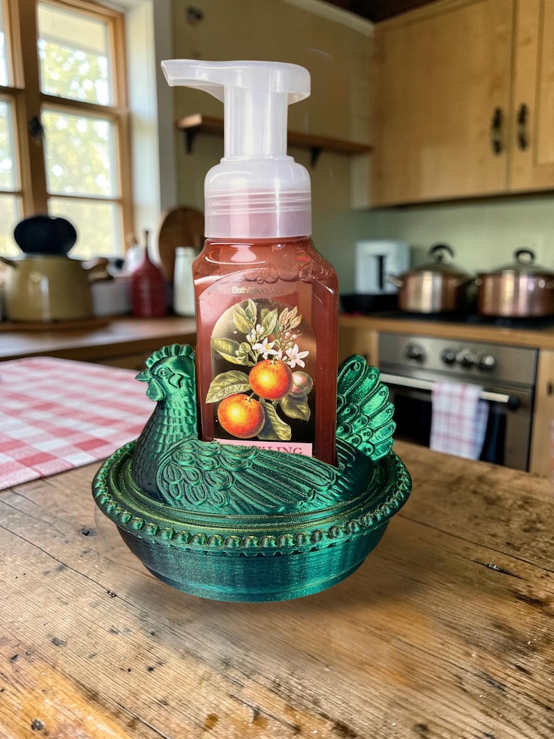 以下が含まれることがあります： 緑色の鶏の形をした石鹸ホルダーと石鹸のボトル。石鹸のボトルには、オレンジと白い花の画像が描かれたラベルが付いています。ホルダーは木製の表面にあります。
