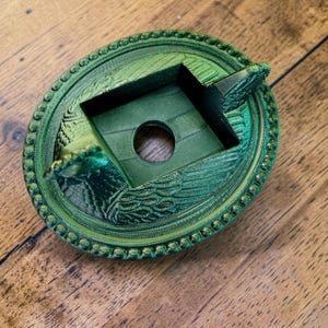 以下が含まれることがあります： 緑色の3Dプリント灰皿。装飾的な円形デザインで、中央に穴のある正方形のエリアがあり、詳細な縁取りと装飾要素で囲まれています。色は鮮やかなメタリックグリーンです。
