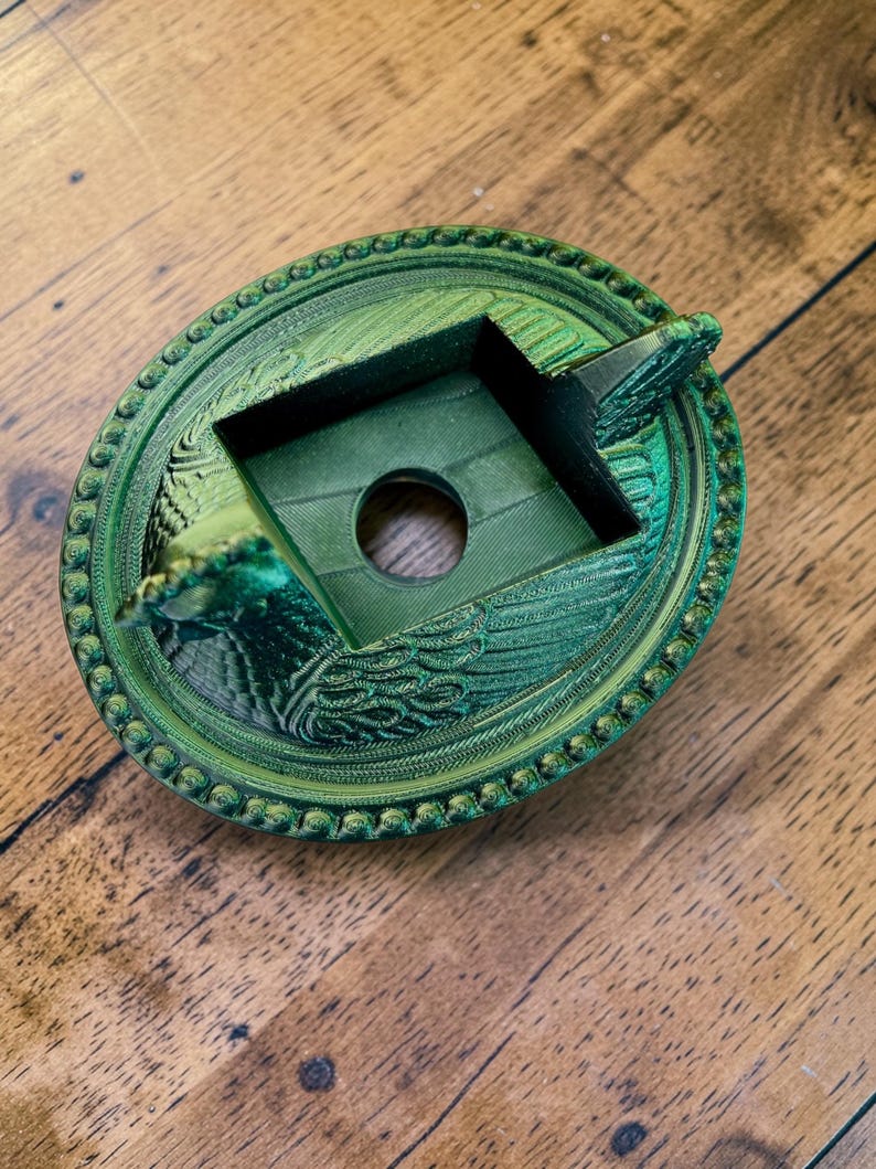 以下が含まれることがあります： 緑色の3Dプリント灰皿。装飾的な円形デザインで、中央に穴のある正方形のエリアがあり、詳細な縁取りと装飾要素で囲まれています。色は鮮やかなメタリックグリーンです。