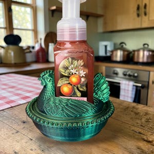 以下が含まれることがあります： 石鹸のボトルを保持する鶏の形をした緑色の装飾石鹸皿。石鹸のボトルはオレンジ色で、白いポンプと果物のイラストが描かれたラベルが付いています。お皿は木製の表面にあります。