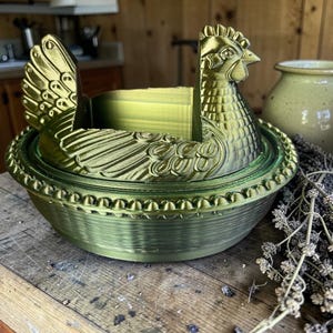 以下が含まれることがあります： メタリックグリーンの鶏の形をした装飾容器。容器は、翼ととさかを持つ鶏の詳細なデザインが特徴です。木製の表面に置かれています。背景には陶器の瓶とドライフラワーがあります。