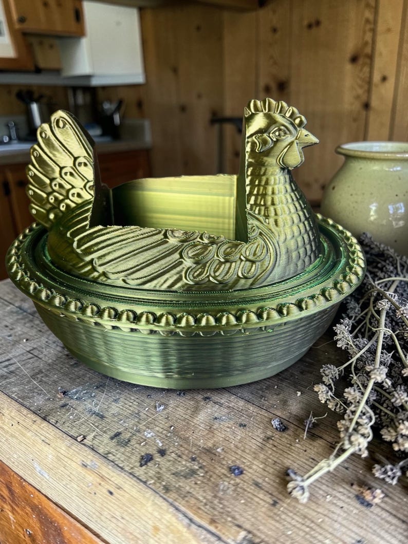 以下が含まれることがあります： メタリックグリーンの鶏の形をした装飾容器。容器は、翼ととさかを持つ鶏の詳細なデザインが特徴です。木製の表面に置かれています。背景には陶器の瓶とドライフラワーがあります。