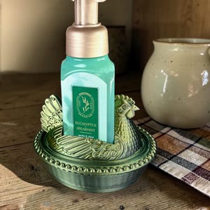 以下が含まれることがあります： 金色のポンプが付いた緑色の石鹸ディスペンサーが、鶏の形をした装飾的な緑色のホルダーに置かれています。石鹸ボトルには、ターコイズ色の液体と「Eucalyptus Spearmint」と書かれたラベルが付いています。ホルダーは木製の表面にあります。