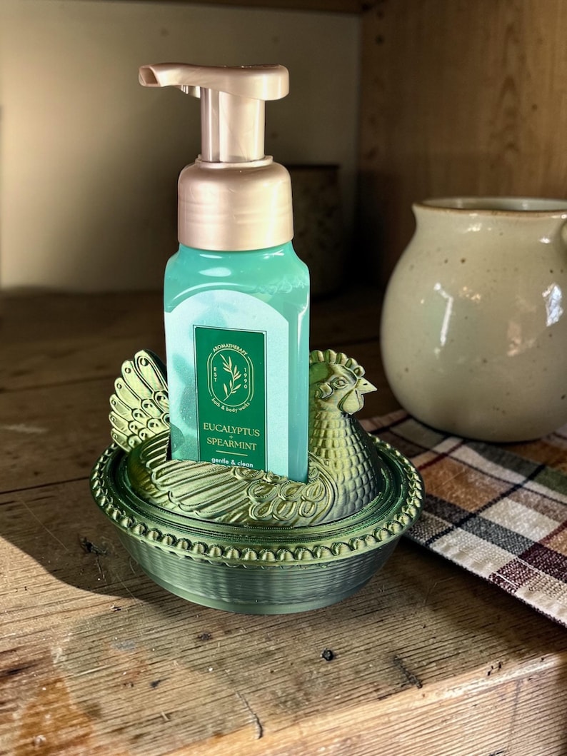 以下が含まれることがあります： 金色のポンプが付いた緑色の石鹸ディスペンサーが、鶏の形をした装飾的な緑色のホルダーに置かれています。石鹸ボトルには、ターコイズ色の液体と「Eucalyptus Spearmint」と書かれたラベルが付いています。ホルダーは木製の表面にあります。