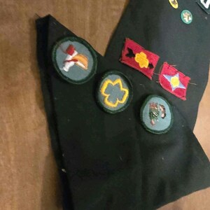 May include: Dark green fabric with various embroidered patches. Patches include a red and yellow flower, a blue star, and a bird. Additional patches are small and circular, with gold and green detailing.