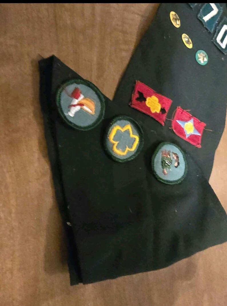May include: Dark green fabric with various embroidered patches. Patches include a red and yellow flower, a blue star, and a bird. Additional patches are small and circular, with gold and green detailing.