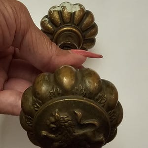 May include: Two antique brass door knobs. One knob has a fluted design with white paint remnants. The other knob has a circular design with a lion emblem. These vintage knobs are likely from a cabinet or door.