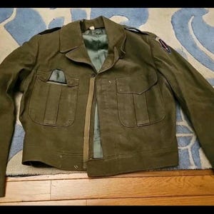May include: Olive green military jacket with two chest pockets and two lower pockets. The jacket has a button closure and a collar. Patches are visible on the sleeves and shoulders. The jacket is laid out on a patterned rug.