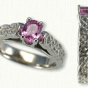 May include: A silver Celtic knot engagement ring with a pink oval-shaped gemstone. The ring is set with a smaller pink gemstone on each side of the main stone.