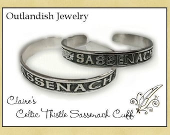 May include: Two silver bracelets with the word "Sassenach" engraved on them. The bracelets are stacked on top of each other. The bracelets are part of a set called "Claire's Celtic Thistle Sassenach Cuff".