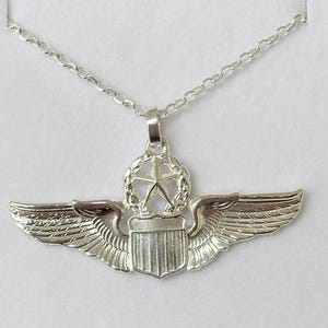 May include: Silver-toned pendant necklace featuring a detailed pilot's wings design. The pendant includes a central star within a wreath, a shield, and stylized wings. The chain is a delicate link style.