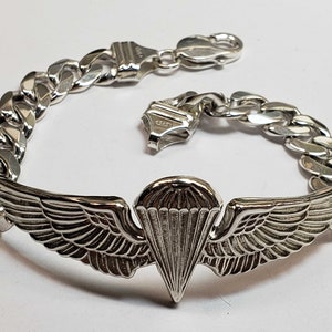 May include: A silver bracelet with a detailed parachute and wings charm. The bracelet is made of a thick, curb chain style.
