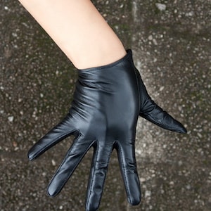 May include: A black leather glove with a smooth, matte finish and visible stitching along the fingers. The glove features a short, straight-cut wrist opening and a fitted design that contours to the shape of the hand, showcasing natural creases.
