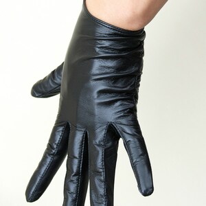 May include: A black leather glove with a smooth, slightly reflective finish and visible stitching along the fingers. The glove features a fitted wrist opening and a soft, pliable texture that conforms to the shape of the hand, showing natural creases.