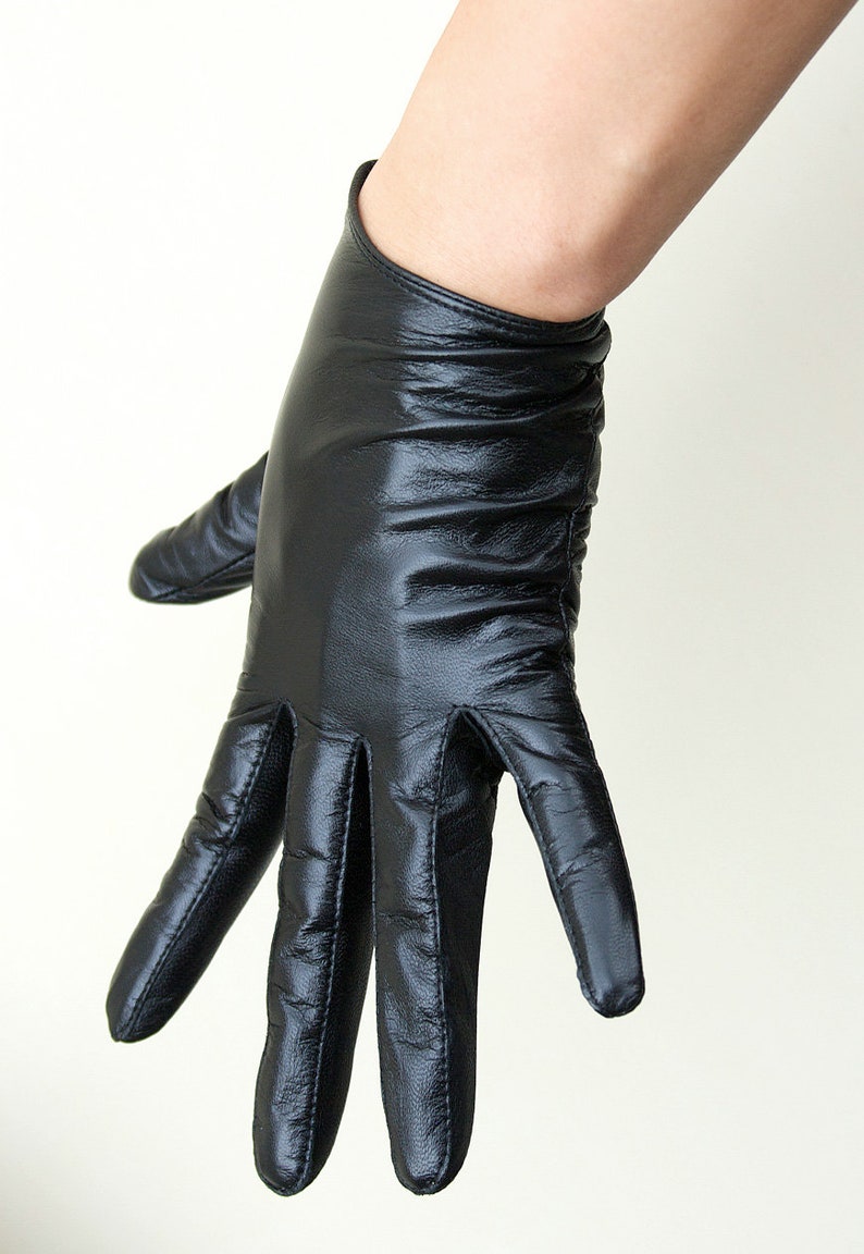 May include: A black leather glove with a smooth, slightly reflective finish and visible stitching along the fingers. The glove features a fitted wrist opening and a soft, pliable texture that conforms to the shape of the hand, showing natural creases.