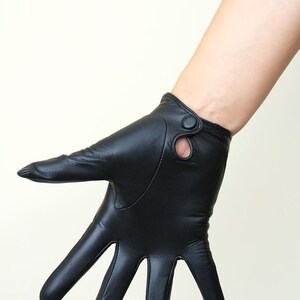 May include: A black leather glove featuring a smooth finish and a wrist strap detail with a small circular cutout and a matching black button closure. The glove covers the fingers and palm, extending slightly past the wrist with a clean seam.