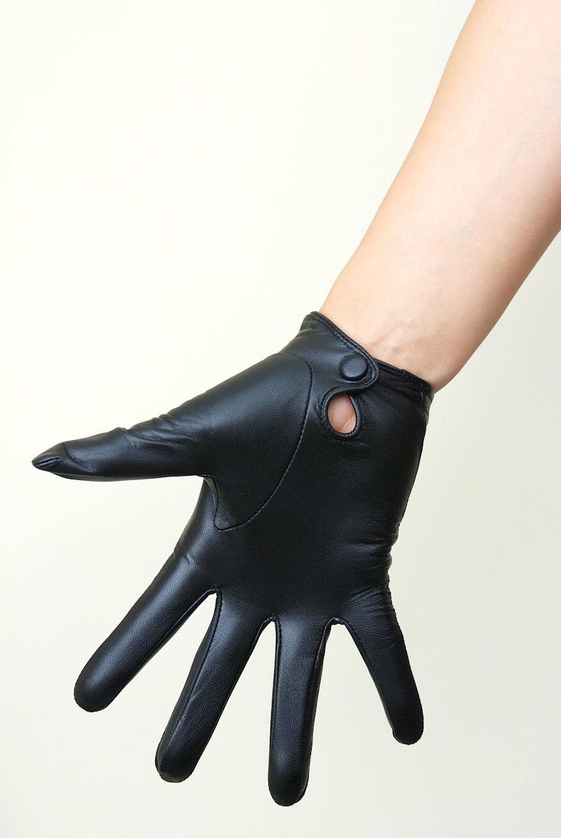 May include: A black leather glove featuring a smooth finish and a wrist strap detail with a small circular cutout and a matching black button closure. The glove covers the fingers and palm, extending slightly past the wrist with a clean seam.