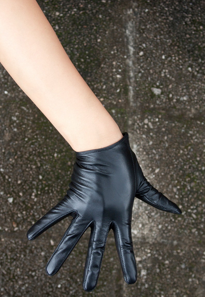 May include: A black leather glove with a smooth, matte finish and visible stitching along the fingers. The glove features a short, straight-cut wrist opening and a fitted design that contours to the shape of the hand, showcasing natural creases.