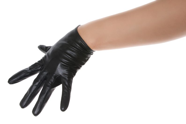 May include: A black leather glove with a smooth, slightly shiny finish and subtle horizontal folds around the wrist area. The glove features stitched seams along the fingers and palm, extending to cover the hand and wrist with a fitted design.