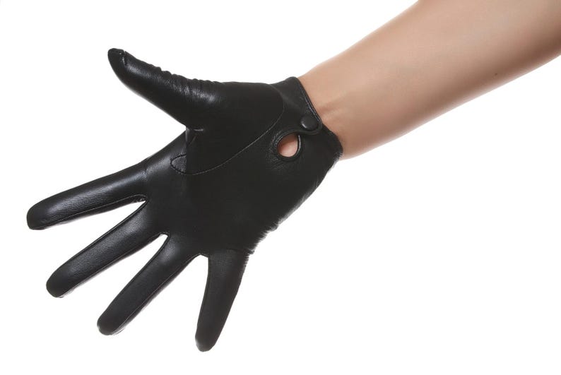 May include: A black leather glove featuring a smooth finish, five individual finger sections, and a small circular cutout detail near the wrist area secured with a single matching black snap button closure for a secure fit around the hand.