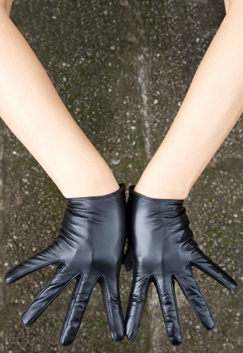 May include: A pair of black leather gloves with a smooth, polished finish and visible stitching along the fingers. The gloves extend to the wrist and feature a soft, flexible texture that conforms to the shape of the hands, showing natural creases.