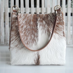May include: A brown and white cowhide tote bag with brown leather straps and gold metal hardware.