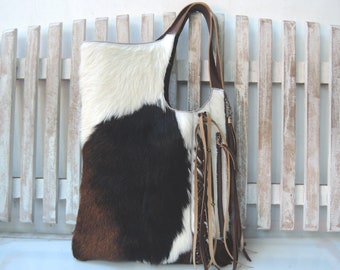 May include: A tote bag crafted from cowhide featuring a tri-color pattern of white, black, and brown patches. The bag includes a single shoulder strap, a vertical zippered pocket on the front, and decorative leather fringe accents hanging down.
