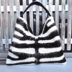 May include: A hobo-style handbag featuring a black and white zebra-striped pattern with a textured hair-on-hide finish. The bag has a single black shoulder strap and a wide, curved opening at the top, showcasing a symmetrical striped design.