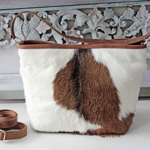 May include: A brown and white cowhide tote bag with a brown leather strap. The bag has a brown leather trim around the top.