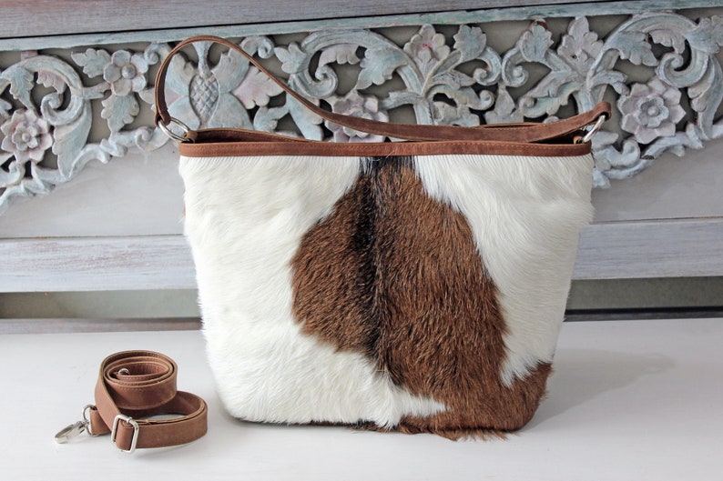May include: A brown and white cowhide tote bag with a brown leather strap. The bag has a brown leather trim around the top.