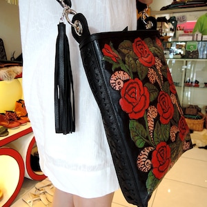 May include: A black leather shoulder bag with a floral design featuring red roses and green leaves. The bag has a long strap and a tassel.